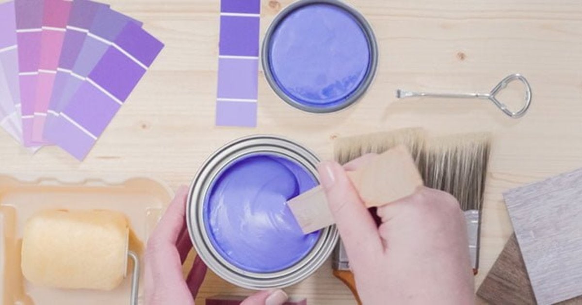 Two hands stirring a tin of lilac paint with a wooden paddle, next to paint swatch cards, a brush and a roller tray
