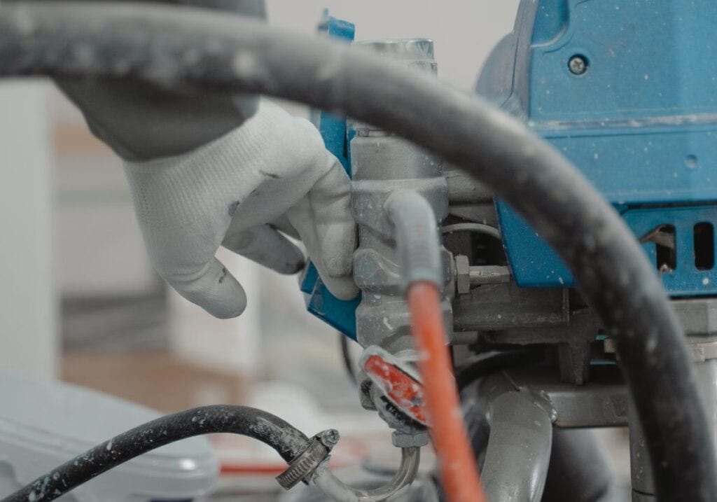 Gloved hands at the pump and suction hose of an airless sprayer, where pattern faults are mechanical more often than mix-related.